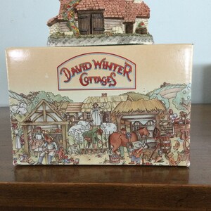 Puede incluir: Una figura decorativa de una casa de campo con tejado de tejas rojas y paredes de piedra se encuentra sobre una caja. La caja presenta el texto "David Winter Cottages" y una ilustración detallada de una escena de pueblo.
