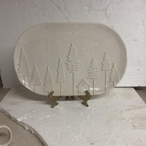 May include: An off-white, oval-shaped ceramic platter with a raised relief design of a winter forest scene. The design includes stylized trees, a small house, and stars. The platter is displayed on a gold-colored metal stand.