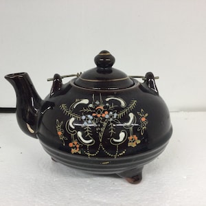 May include: A dark brown ceramic teapot with a lid and a spout. The teapot has a decorative floral design in white, orange, and blue. It has a metal handle and sits on three small feet. The teapot is likely vintage or antique.