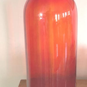 May include: A tall, cylindrical glass vase featuring a vibrant, translucent red hue. The vessel has a smooth, polished surface and a wide, flared rim that tapers into a slender neck before widening into a straight-sided body with a flat base.