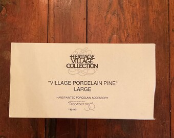 May include: A rectangular box with the text "Heritage Village Collection" and "Village Porcelain Pine" in black. The box also says "Handpainted Porcelain Accessory" and "Large". The box is on a wooden surface.