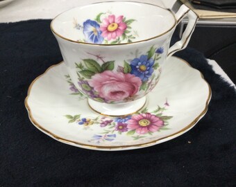 May include: A white teacup and saucer set with a floral design. The teacup and saucer have a scalloped edge and gold trim. The floral design includes pink, blue, and purple flowers with green leaves.