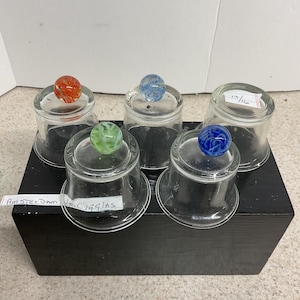 May include: Five clear glass containers with colorful marble-like toppers. The marbles are red, blue, green, and a mix of colors. The containers are on a black wooden box. Text on a label reads "Amsterdam No. C 1991AS".