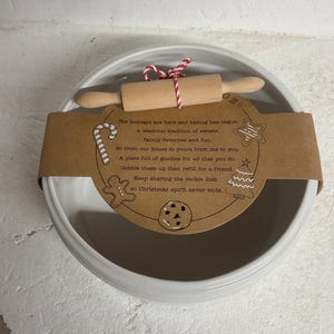 May include: A white ceramic cookie dish with a small wooden rolling pin tied with red and white twine. A brown paper label with holiday-themed illustrations and text is wrapped around the dish.