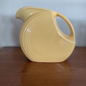 May include: A pale yellow ceramic pitcher with a curved handle and a spout. The pitcher has a rounded body with concentric circular ridges. The pitcher is sitting on a wooden surface.