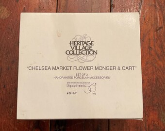 May include: A rectangular box with the text "The Heritage Village Collection" and "Chelsea Market Flower Monger & Cart". The box also includes the text "Set of 2 Handpainted Porcelain Accessories" and the Department 56 logo.