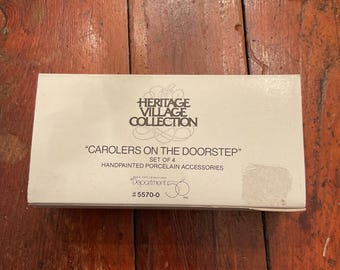 May include: A white box from the Heritage Village Collection, featuring the text "CAROLERS ON THE DOORSTEP" and "HANDPAINTED PORCELAIN ACCESSORIES". The box is set on a wooden surface.