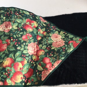 K&ouml;nnte beinhalten: Ein dekoratives Textil mit dunkelgr&uuml;nem Hintergrund, das ein Blumenmuster aus roten &Auml;pfeln, rosa Rosen und roten Beeren zeigt. Der Stoff hat einen gr&uuml;n gen&auml;hten Rand und bedeckt teilweise eine schwarze, strukturierte Oberfl&auml;che.