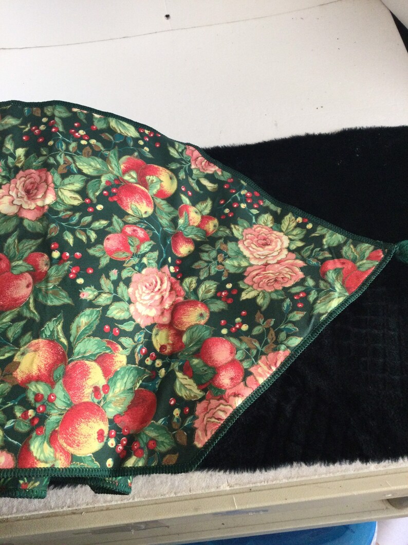 K&ouml;nnte beinhalten: Ein dekoratives Textil mit dunkelgr&uuml;nem Hintergrund, das ein Blumenmuster aus roten &Auml;pfeln, rosa Rosen und roten Beeren zeigt. Der Stoff hat einen gr&uuml;n gen&auml;hten Rand und bedeckt teilweise eine schwarze, strukturierte Oberfl&auml;che.