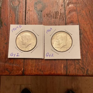 Puede incluir: Dos monedas de medio dólar Kennedy de 1964 en protectores. Las monedas plateadas muestran un perfil de John F. Kennedy. Cada protector está etiquetado con el año y una calificación escrita a mano. Las monedas están sobre una superficie de madera.