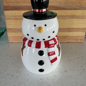 May include: A white snowman-shaped ceramic jar with a black top hat and a red and white striped scarf. The snowman has a yellow carrot nose, black button eyes, and three black buttons down the front. A festive holiday decoration.