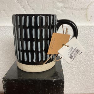 May include: A black stoneware mug with a white vertical dash pattern and a beige base. The mug has a black handle and a tag attached with twine. The tag includes the text "mudpie" and "LINES STONEWARE MUG".