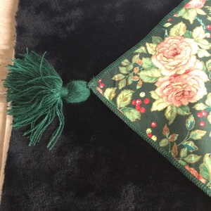K&ouml;nnte beinhalten: Ein schwarzer, pl&uuml;schiger Stoff mit einer gr&uuml;nen Quaste und einer Ecke mit Blumenmuster. Die Ecke zeigt rosa Rosen, rote Beeren und gr&uuml;ne Bl&auml;tter auf einem dunkelgr&uuml;nen Hintergrund. Die Quaste besteht aus gr&uuml;nem Garn.