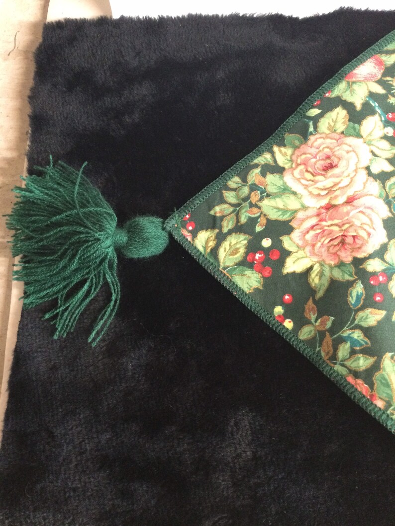 K&ouml;nnte beinhalten: Ein schwarzer, pl&uuml;schiger Stoff mit einer gr&uuml;nen Quaste und einer Ecke mit Blumenmuster. Die Ecke zeigt rosa Rosen, rote Beeren und gr&uuml;ne Bl&auml;tter auf einem dunkelgr&uuml;nen Hintergrund. Die Quaste besteht aus gr&uuml;nem Garn.