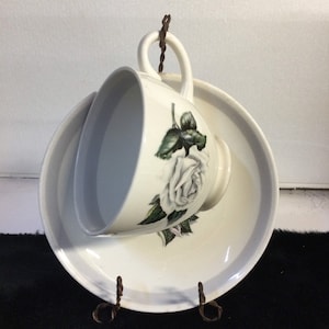 May include: A white teacup and saucer set with a floral rose design. The teacup and saucer are displayed on a decorative metal stand. The teacup features a white rose with green leaves. The set is a vintage style.