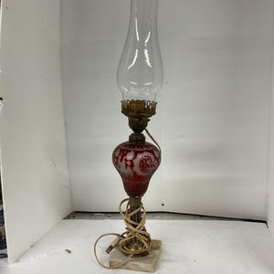 May include: An antique electric lamp with a clear glass chimney and a red glass base. The base features an etched design and sits on a square marble base. The lamp has a brass-colored metal frame and a power cord.
