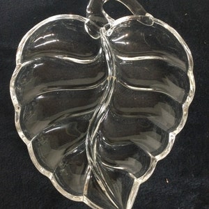 May include: Clear glass leaf-shaped serving dish with a handle. The dish is divided into two sections with a leaf vein design. The edges are scalloped. The handle is a loop shape.