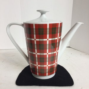 May include: White ceramic teapot with a red, green, and white plaid pattern. The teapot has a curved handle, a long spout, and a lid with a small knob. The teapot is sitting on a black felt coaster.