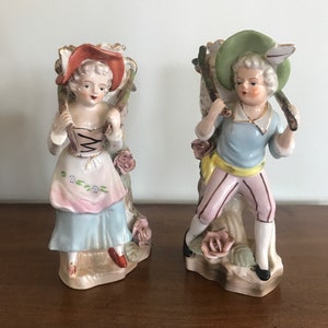 May include: A pair of porcelain figurines depicting a woman and a man in 18th-century attire. The woman wears a pink and blue dress with a red hat. The man carries a bundle on his back and wears a blue shirt and pink pants. Both figures are adorned with floral accents.