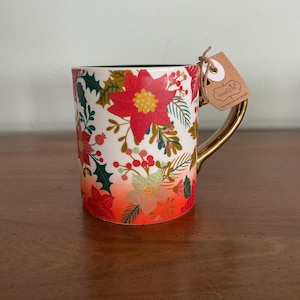 May include: A ceramic mug with a gold handle and a festive floral design. The mug features red poinsettias, green holly, and berries against a white background with an orange gradient. A brown tag is attached to the handle.
