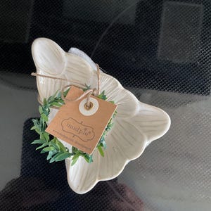 May include: A cream-colored, petal-shaped dish with a small green wreath and a brown tag tied with twine. The tag has the brand name "mudpie" and a website address printed on it.