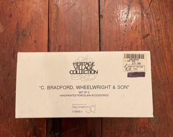 May include: A white box from the Heritage Village Collection, featuring the text "C. Bradford, Wheelwright & Son" and "Handpainted Porcelain Accessories." The box has a price tag and is set on a wooden surface.