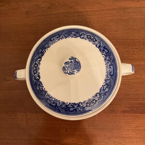 May include: A white and blue ceramic serving dish with a lid. The dish has a blue floral pattern around the rim and on the lid. The handles are white with blue accents.