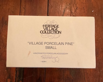 May include: A rectangular box with the text "HERITAGE VILLAGE COLLECTION" and "VILLAGE PORCELAIN PINE" SMALL. The box also reads "HANDPAINTED PORCELAIN ACCESSORY" and "MADE EXCLUSIVELY FOR Department 56". The box is on a wooden surface.
