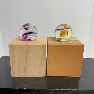 May include: Two glass marbles with colorful swirls sit atop wooden blocks. One marble features purple, black, and white designs, while the other has yellow, orange, and blue patterns. The marbles are clear and spherical.