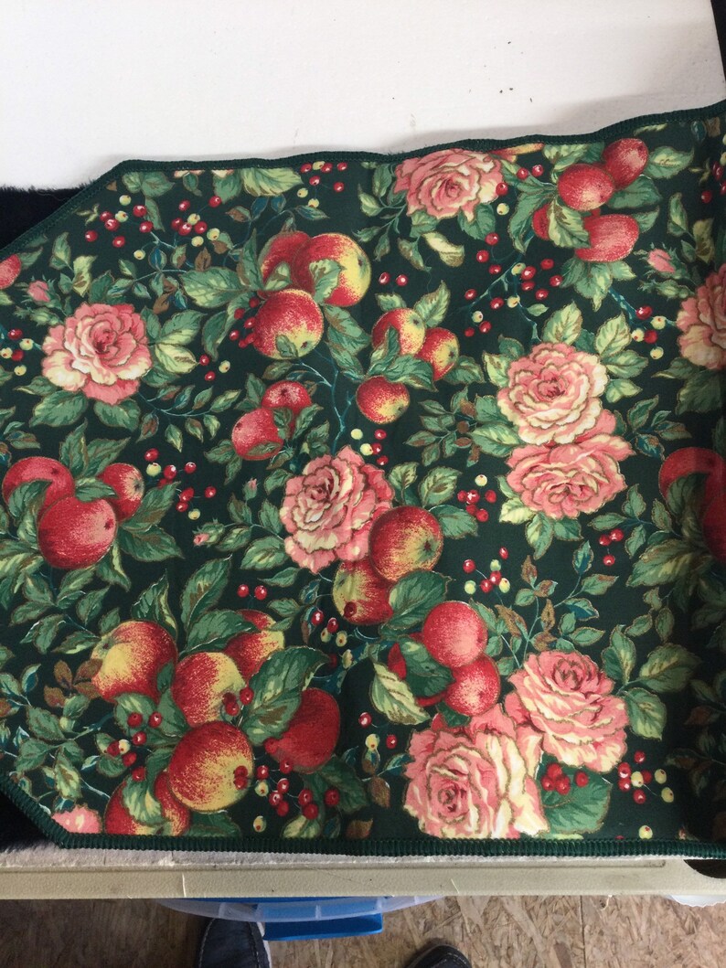 K&ouml;nnte beinhalten: Ein rechteckiger Stoff mit dunkelgr&uuml;nem Hintergrund, der ein Muster aus roten &Auml;pfeln, rosa Rosen und kleinen roten Beeren aufweist. Die Kanten sind mit einem dunkelgr&uuml;nen Rand versehen.