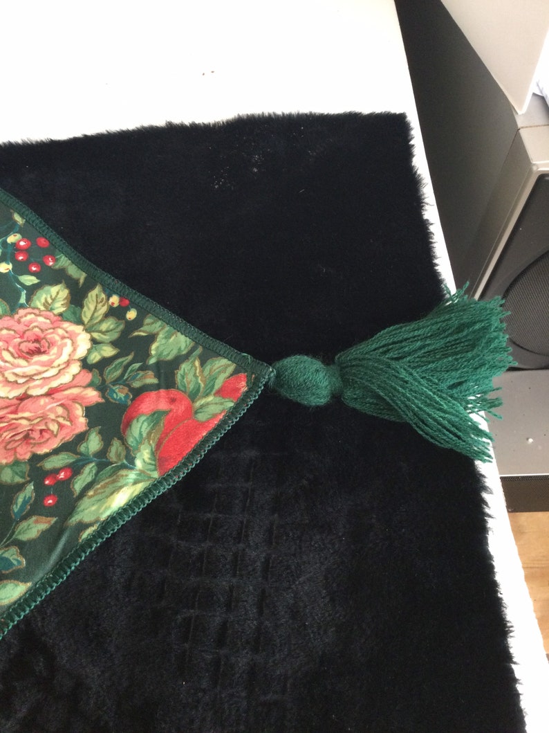 K&ouml;nnte beinhalten: Dekorativer Tischl&auml;ufer mit einem Blumenmuster aus rosa Rosen und roten &Auml;pfeln auf dunkelgr&uuml;nem Grund. Er hat eine gr&uuml;ne Quaste und liegt auf einer schwarzen, strukturierten Oberfl&auml;che. Dekorationsartikel.