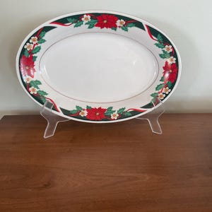 May include: An oval white ceramic serving platter featuring a decorative border of red poinsettia flowers, green holly leaves, and thin red ribbons. The platter is displayed on a clear plastic stand, showcasing its smooth, glossy finish.