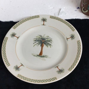 May include: Off-white ceramic plate featuring a central palm tree illustration with a brown trunk and green fronds. The plate has a gold rim and a green and gold geometric border, with smaller palm tree illustrations around the edge.