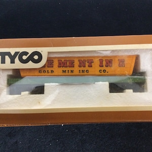 May include: A vintage TYCO model train set in its original packaging. The box is brown with a clear window showing the train car. The train car is orange with the words "Gold Mining Co." printed on the side. The set is a collectible item.