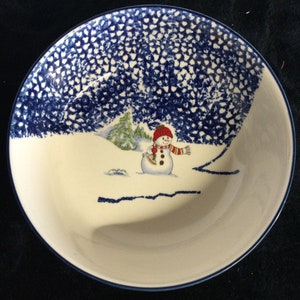 May include: A white ceramic bowl with a blue rim and a winter scene. The bowl features a snowman wearing a red hat and scarf, with a snowy landscape and trees. The rim has a blue dotted pattern.