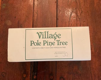 May include: A white box with green text that reads "Village Pole Pine Tree." The box also states "Contents: One 8" Cold Cast Porcelain Tree." The box is on a wooden surface.