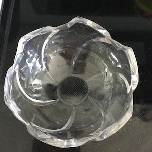 May include: A clear, decorative glass bowl with a flower-like design. The bowl has a circular, textured center and curved, petal-shaped sides. The bowl is made of clear glass and is sitting on a dark surface.