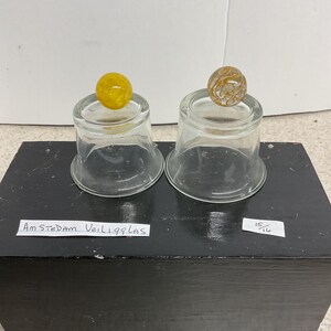May include: Two clear glass candle holders each topped with a decorative marble. One marble is yellow, the other is tan with a swirl pattern. The candle holders sit on a black wooden block. Text on the block reads "Amsterdam Veil 199 Las".