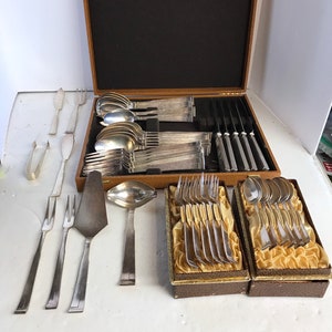 May include: A vintage silver-toned flatware set, including forks, knives, spoons, and serving utensils, displayed in a wooden case with a dark interior. Additional pieces are arranged outside the case, showcasing a complete dining set.