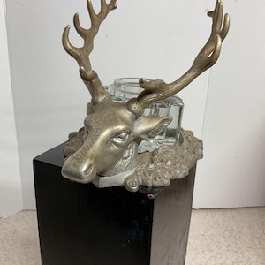 May include: An antique inkwell featuring a deer head design with detailed antlers. The inkwell is made of silver-toned metal and has a clear glass ink pot. It sits atop a black wooden pedestal.