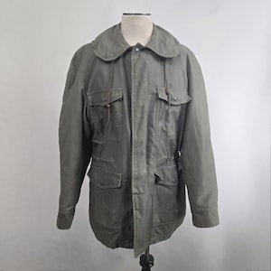 May include: Olive green military-style jacket with a rounded collar and multiple pockets. The jacket has a drawstring waist and button closures. The jacket is displayed on a mannequin.