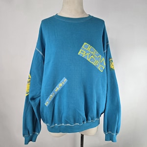 May include: Teal blue sweatshirt with the text "OCEAN PACIFIC" in yellow block letters. The sweatshirt has patches with yellow and blue designs. White stitching accents the seams and cuffs.