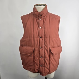 May include: A rust-colored, quilted puffer vest with a high collar and two front pockets. The vest has a button-up closure with silver-toned buttons. The vest is displayed on a mannequin.