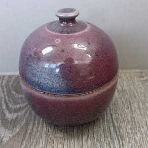 May include: A spherical ceramic jar with a lid, featuring a glossy glaze in shades of deep purple and hints of blue. The surface has a subtle crackle texture and a small, rounded knob handle on top. The container has a smooth, rounded silhouette.