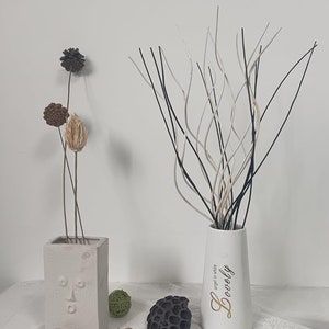 May include: A white ceramic vase with the word "Lovely" written in gold lettering and the phrase "angel in white" in smaller lettering. The vase is filled with black and white decorative branches. The vase is sitting on a white surface with a few decorative items.