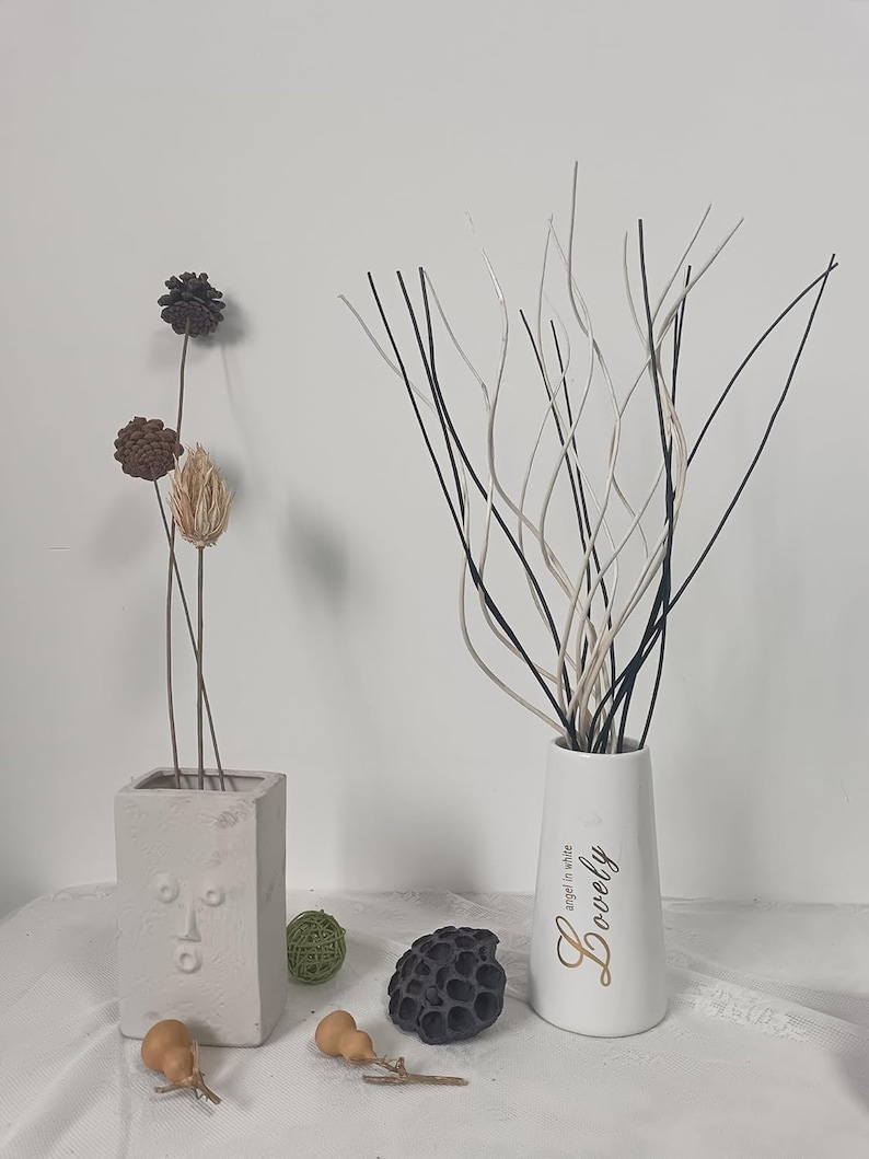 May include: A white ceramic vase with the word "Lovely" written in gold lettering and the phrase "angel in white" in smaller lettering. The vase is filled with black and white decorative branches. The vase is sitting on a white surface with a few decorative items.