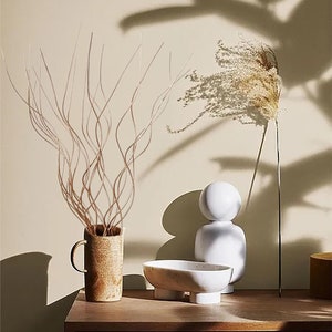 May include: A wooden table with a brown wicker surface. On the table are three decorative objects: a white ceramic sculpture, a white marble bowl with four legs, and a brown ceramic vase with a dried branch arrangement.