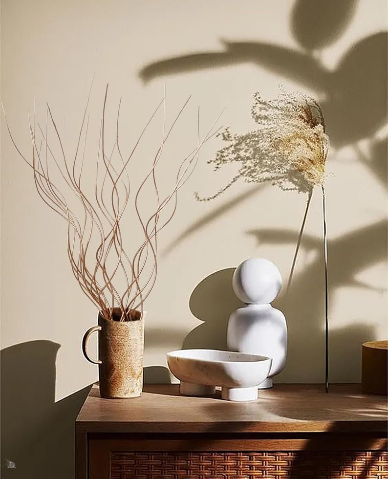 May include: A wooden table with a brown wicker surface. On the table are three decorative objects: a white ceramic sculpture, a white marble bowl with four legs, and a brown ceramic vase with a dried branch arrangement.