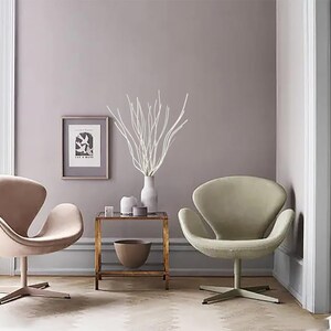 May include: Two white swan chairs in a living room setting with a side table and a vase with white branches. The walls are painted a light purple color.