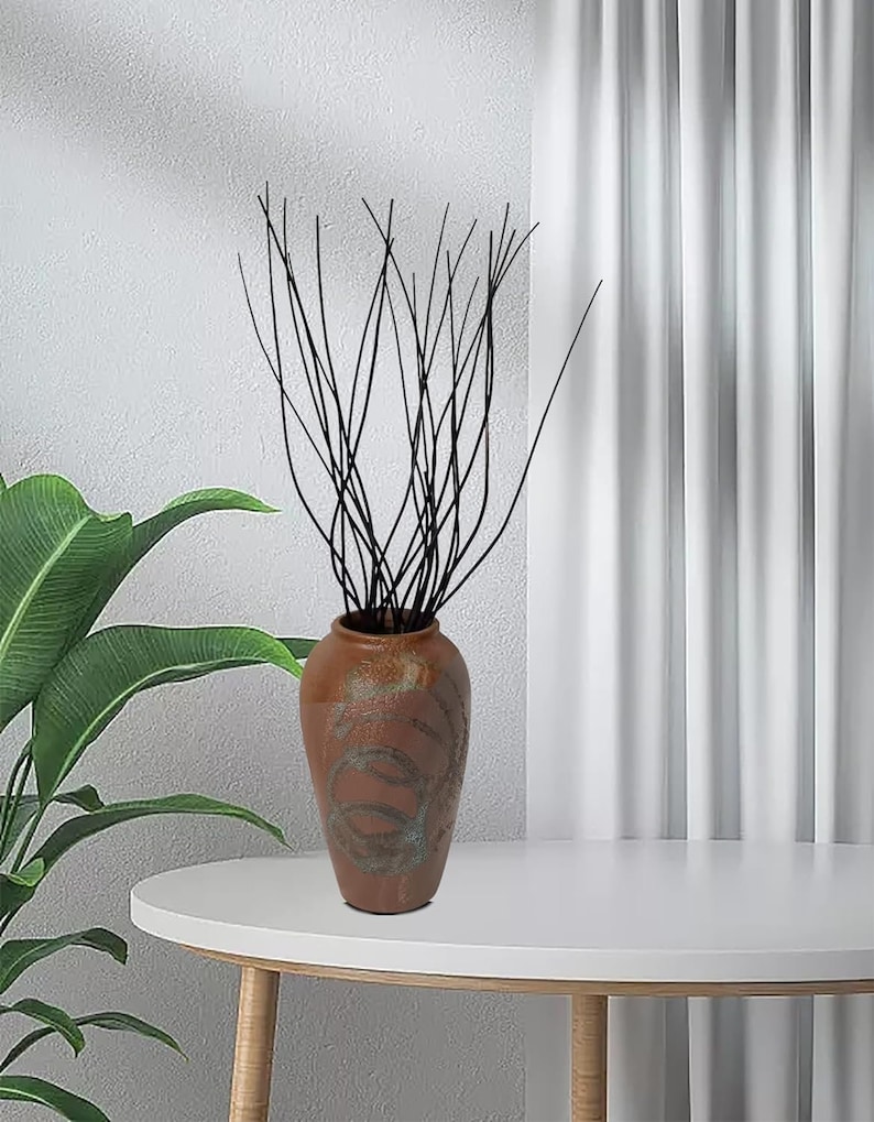 May include: A brown ceramic vase with a swirling pattern, filled with black twig branches. The vase is sitting on a white table.